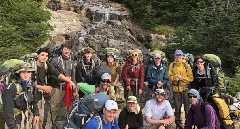 A group of people wearing backpacks pose for a photo in front of a small waterfall.